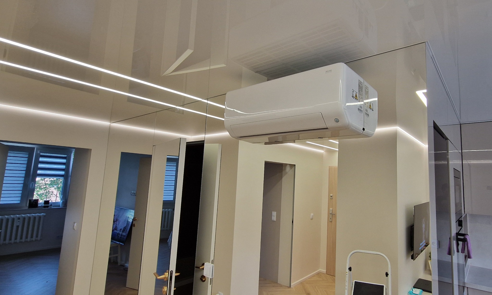 Biała klimatyzacja typu split zamontowana pod sufitem w nowoczesnym wnętrzu z oświetleniem LED, widok z dołu. Na ścianie lustrzane panele.