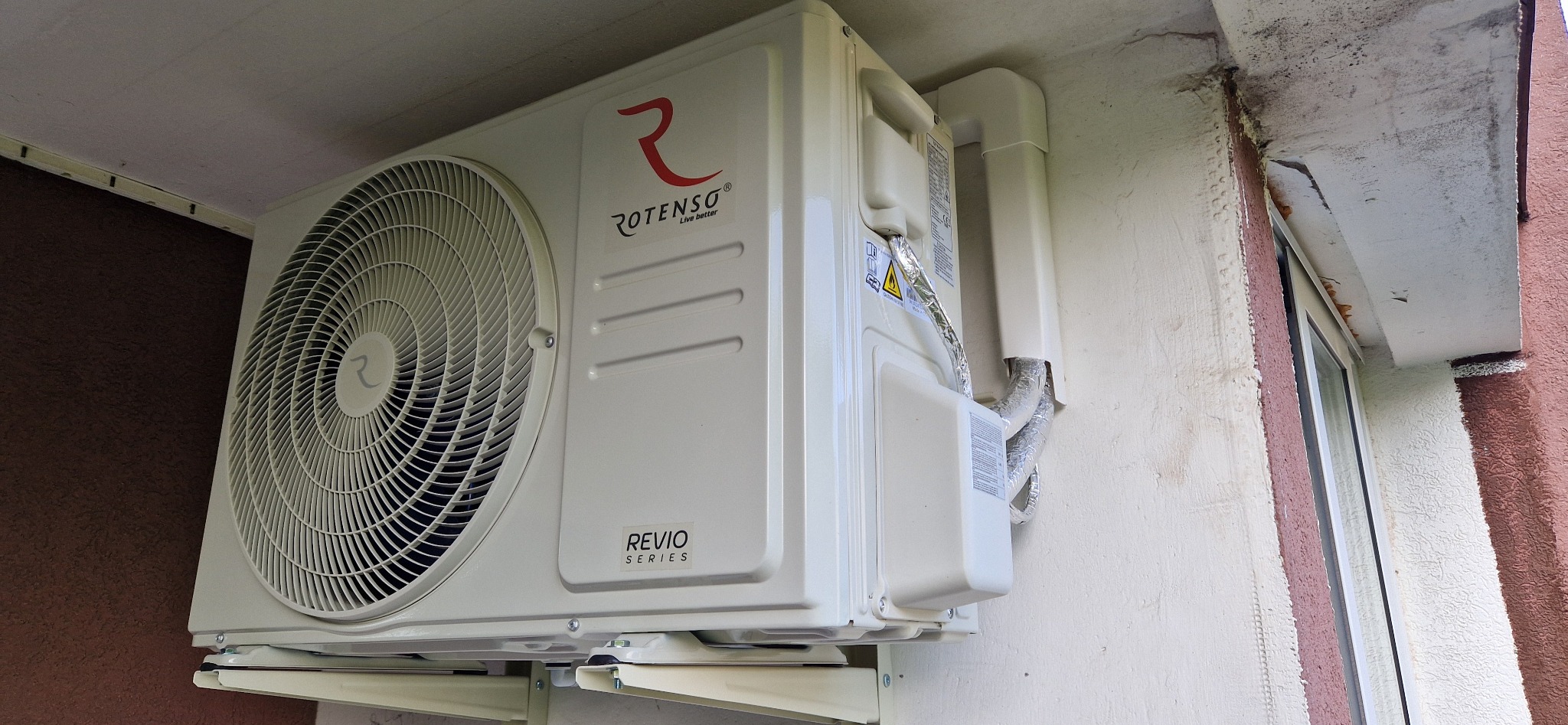 Biały klimatyzator Rotenso Revio Series zamontowany na zewnątrz budynku, widoczne rury odprowadzające, ściana z oknem w tle.