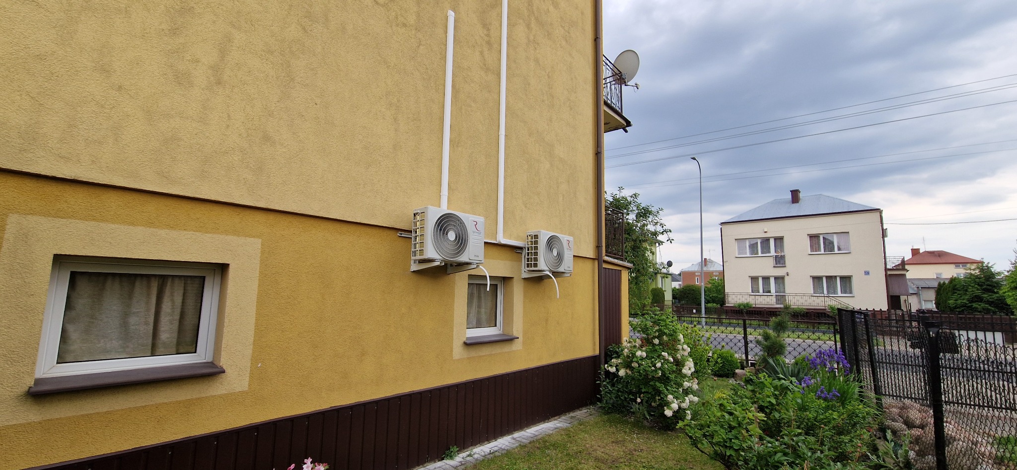 Dwa klimatyzatory na elewacji budynku o żółtym kolorze, z widocznymi białymi rurami odprowadzającymi. W tle zieleń i fragment sąsiedniego budynku.