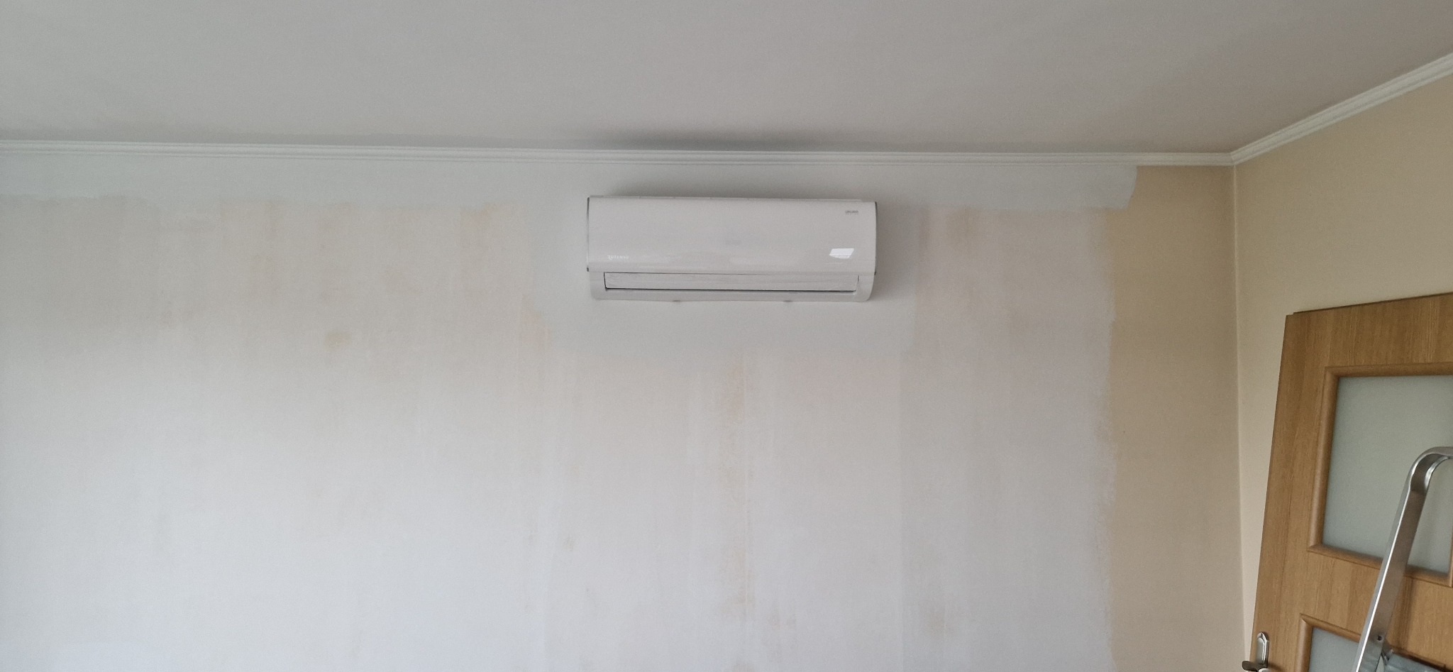 Biały klimatyzator na ścianie w trakcie malowania. Część ściany jest pomalowana na biało, a część w kolorze beżowym. Widoczna drabina oparta o drzwi.