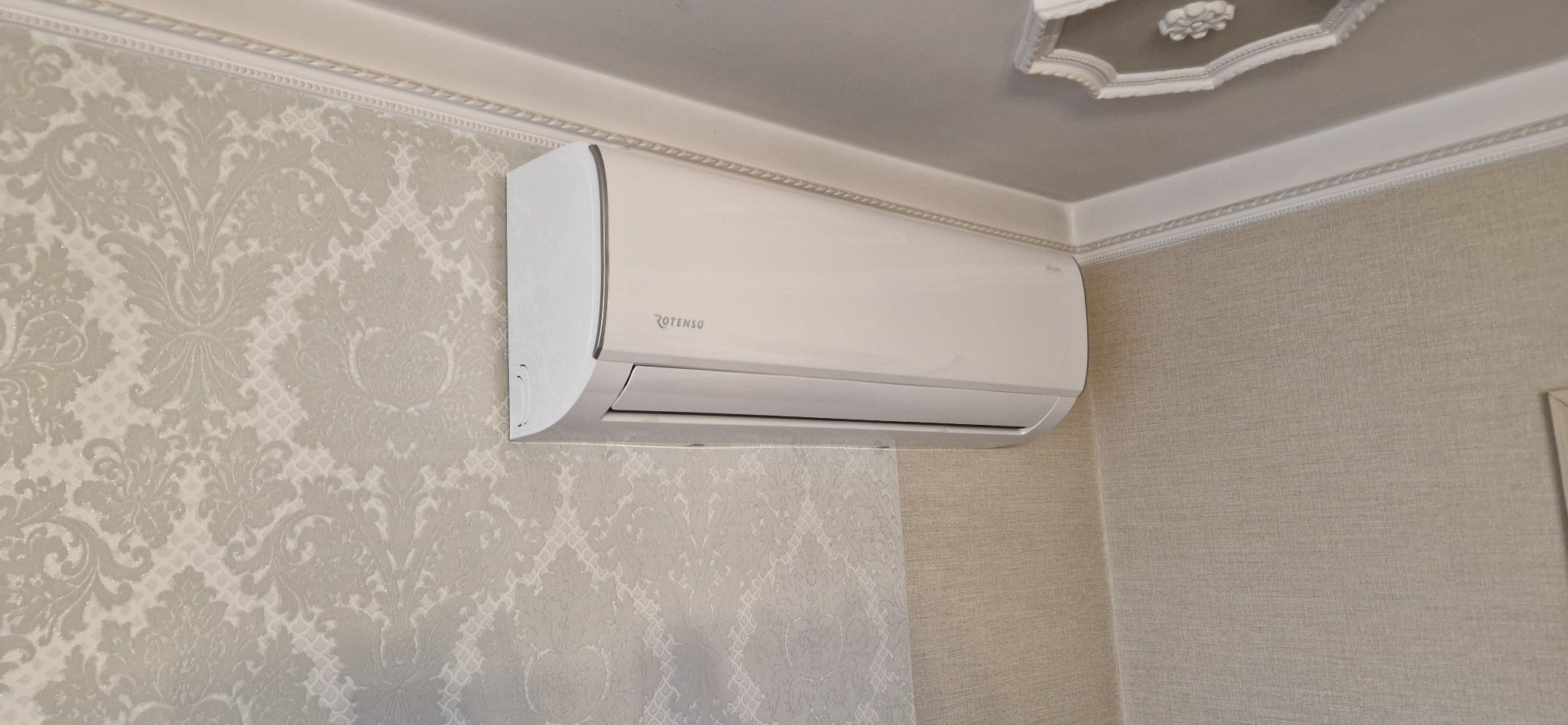 Biała klimatyzacja Rotenso zamontowana na ścianie z wzorzystą tapetą. Widoczne zdobienia sufitowe. Estetyczny montaż w jasnym wnętrzu.