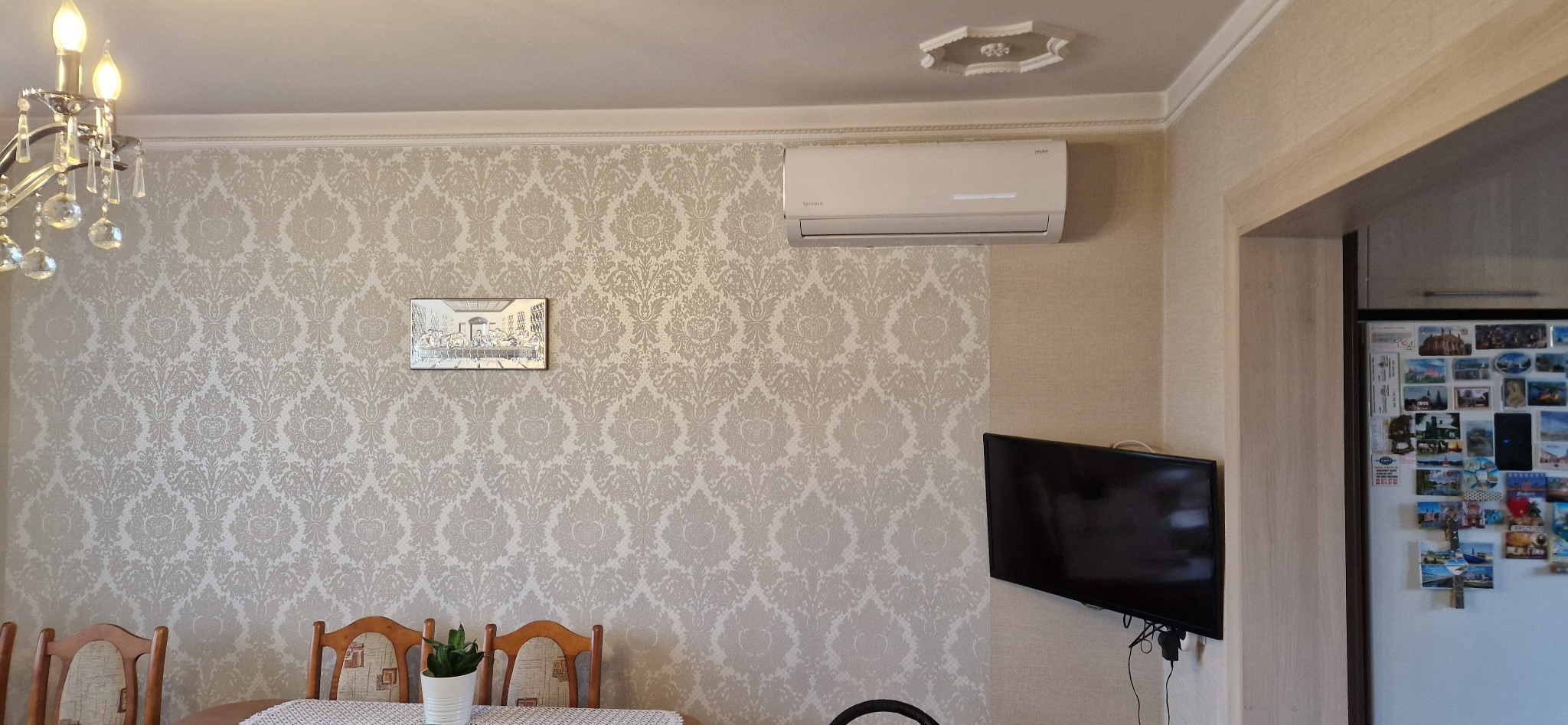 Zamontowany klimatyzator w jasnym wnętrzu pokoju z wzorzystą tapetą, widoczny telewizor i fragment stołu z krzesłami. Widok na lodówkę.