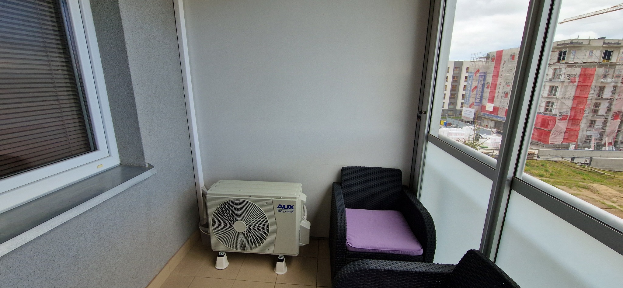 Biały klimatyzator AUX na balkonie mieszkania w bloku. Widoczne czarne fotele z fioletowymi poduszkami i budynek w budowie za oknem.