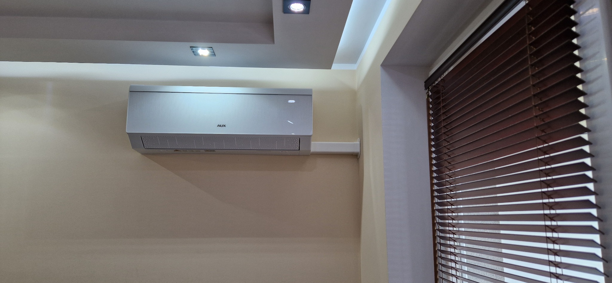 Zamontowana klimatyzacja AUX na ścianie w pomieszczeniu z żaluzjami w oknie. Widoczny fragment sufitu z oświetleniem punktowym.