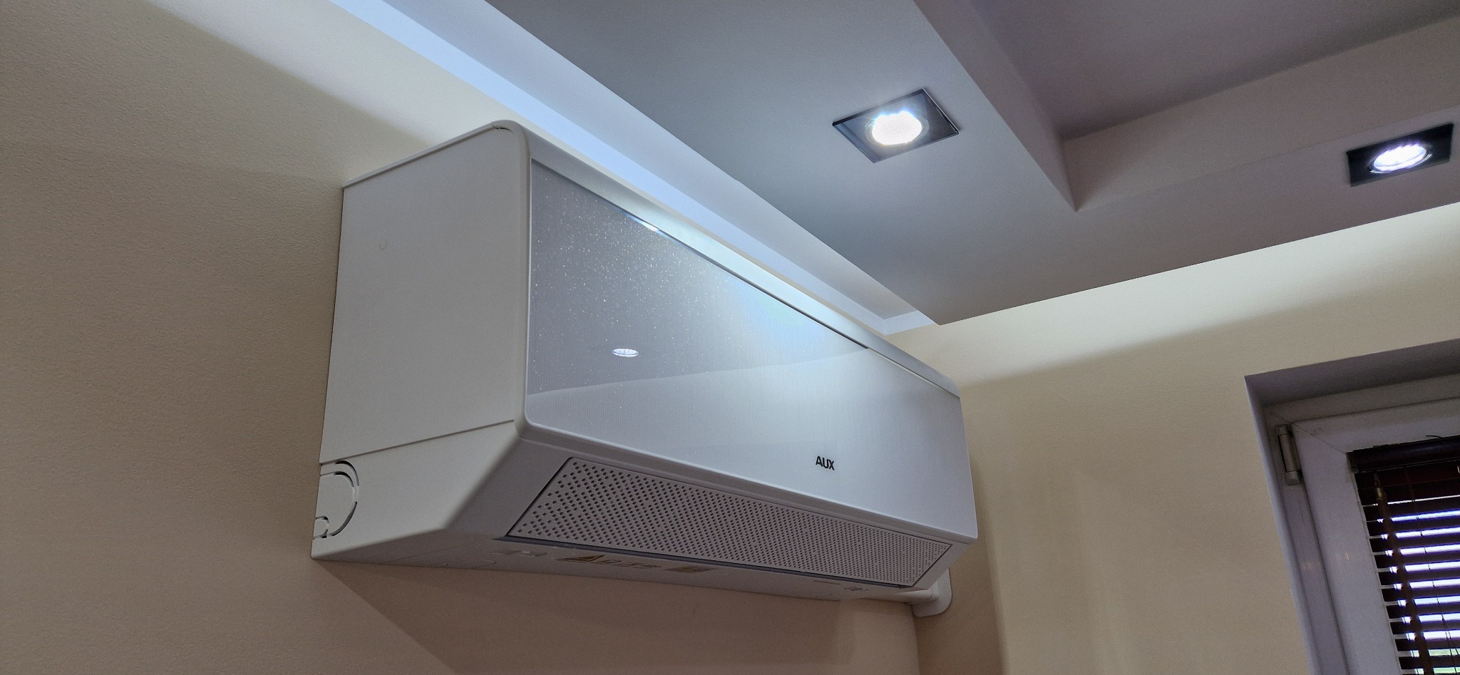 Biały klimatyzator AUX na kremowej ścianie, widoczny z dołu pod kątem. Element wnętrza z oświetleniem sufitowym i fragmentem okna z żaluzjami w tle. Nowoczesny design.