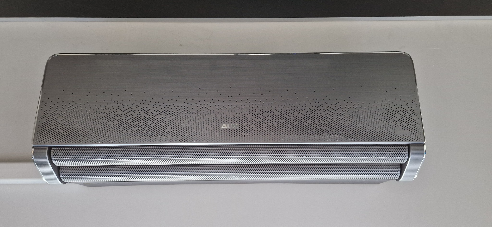 Nowoczesny klimatyzator AUX w metalicznej obudowie z perforowanym wzorem, zamontowany na jasnej ścianie. Widoczne detale wykończenia i perforacja obudowy.