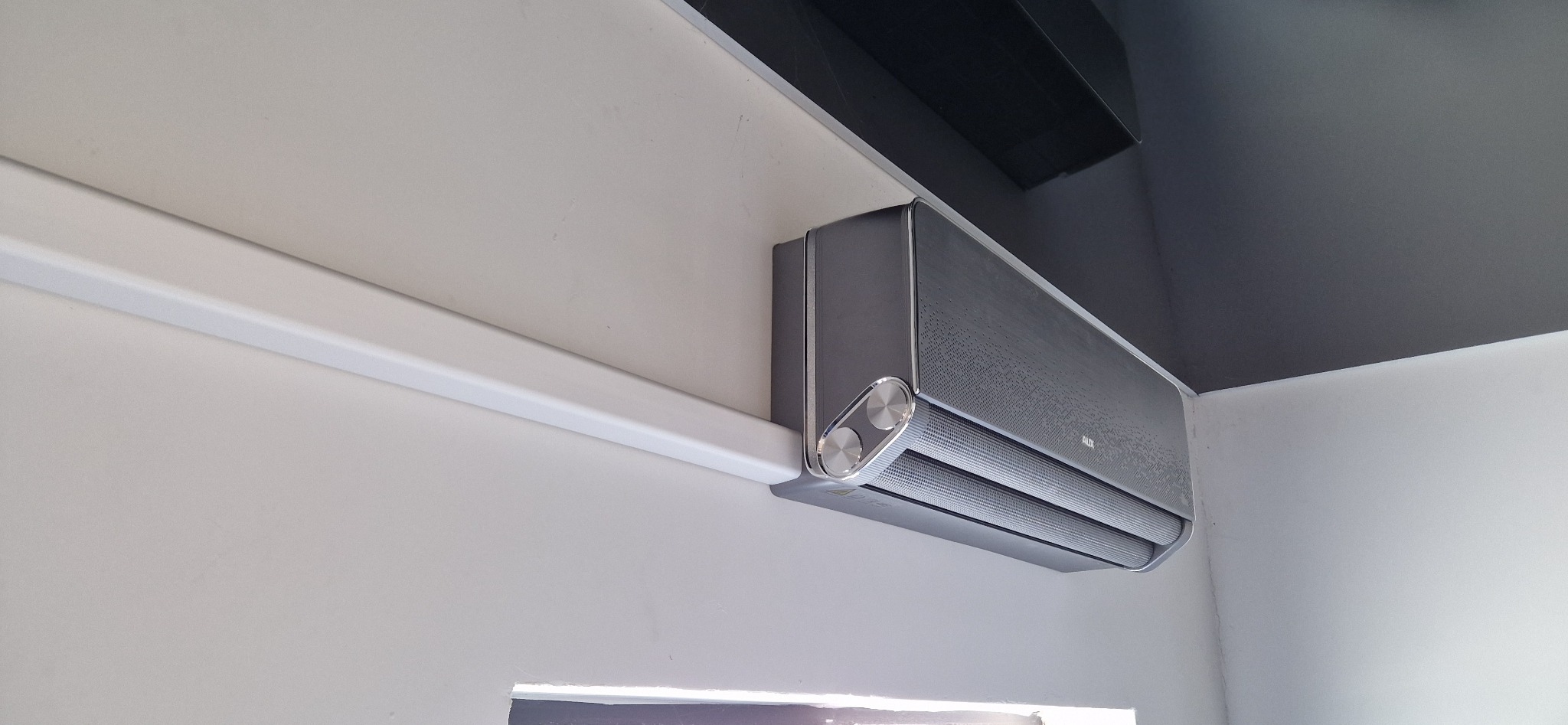 Nowoczesny, szary klimatyzator AUX zamontowany na białej ścianie z widoczną listwą maskującą i fragmentem sufitu w kolorze grafitowym.