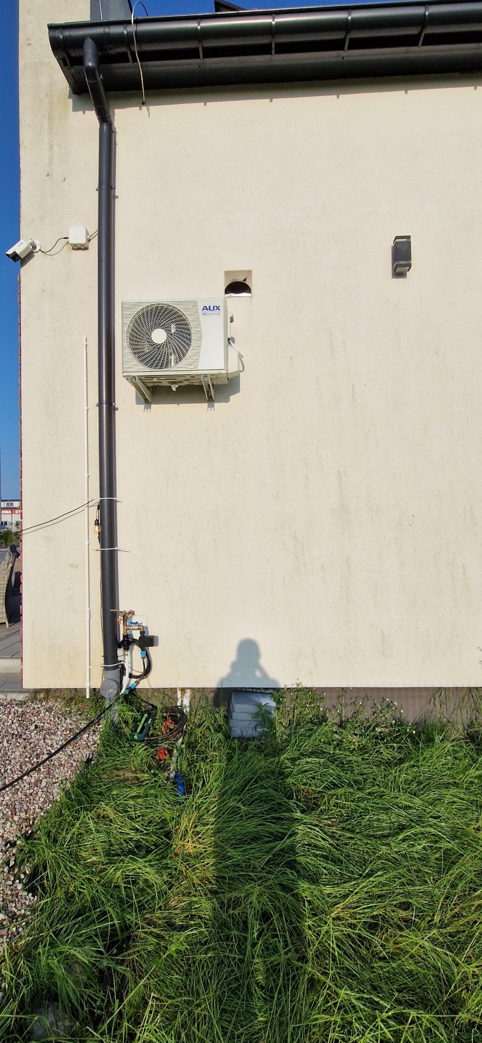 Biała jednostka zewnętrzna klimatyzacji AUX zamontowana na elewacji budynku z widoczną instalacją, rurą spustową i zarośniętą trawą u podstawy ściany.