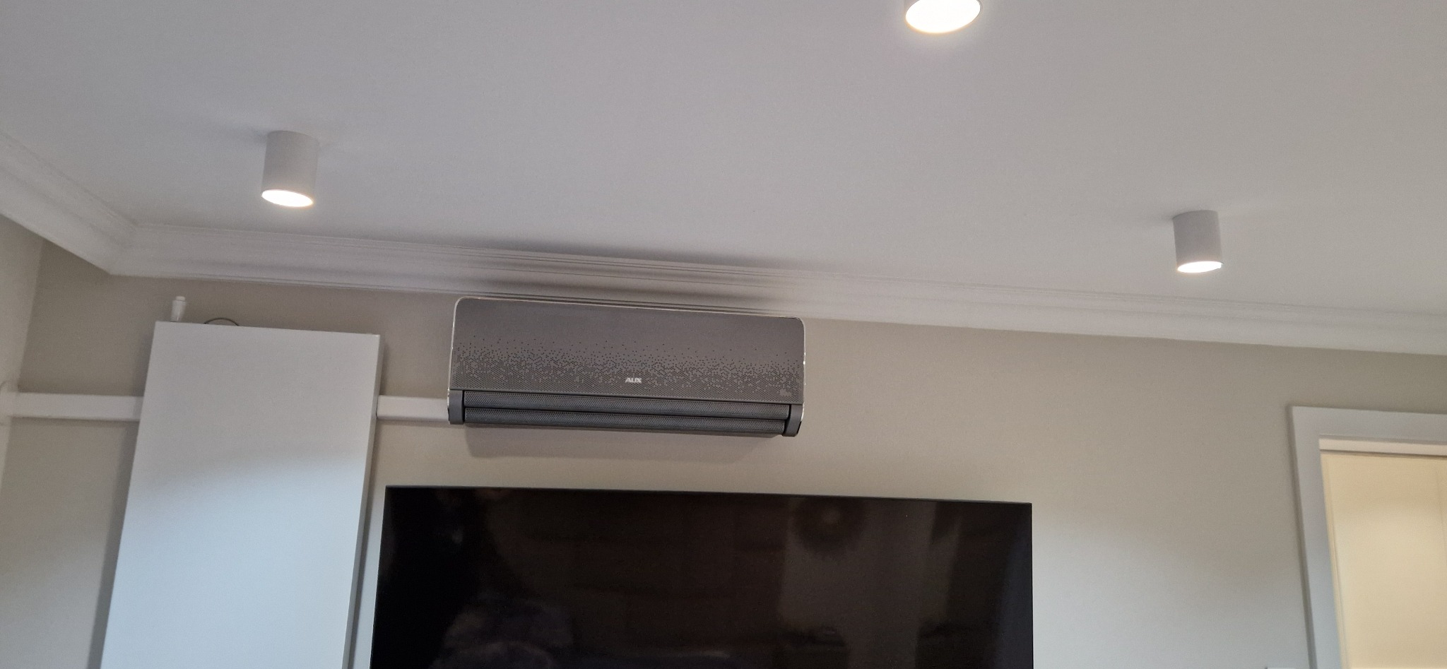 Szara klimatyzacja AUX zamontowana na ścianie w pokoju z podwieszanym sufitem i oświetleniem punktowym, widok z dołu.