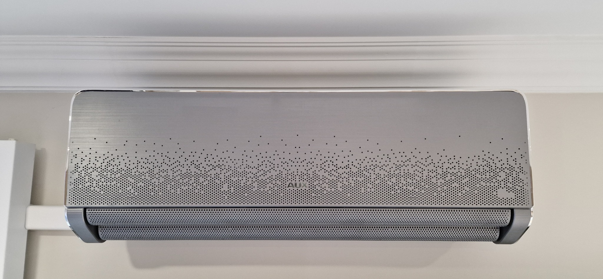 Szary klimatyzator AUX na ścianie, pod sufitem z dekoracyjnym wzorem z kropek. Widoczne detale wykończenia i perforowana obudowa.