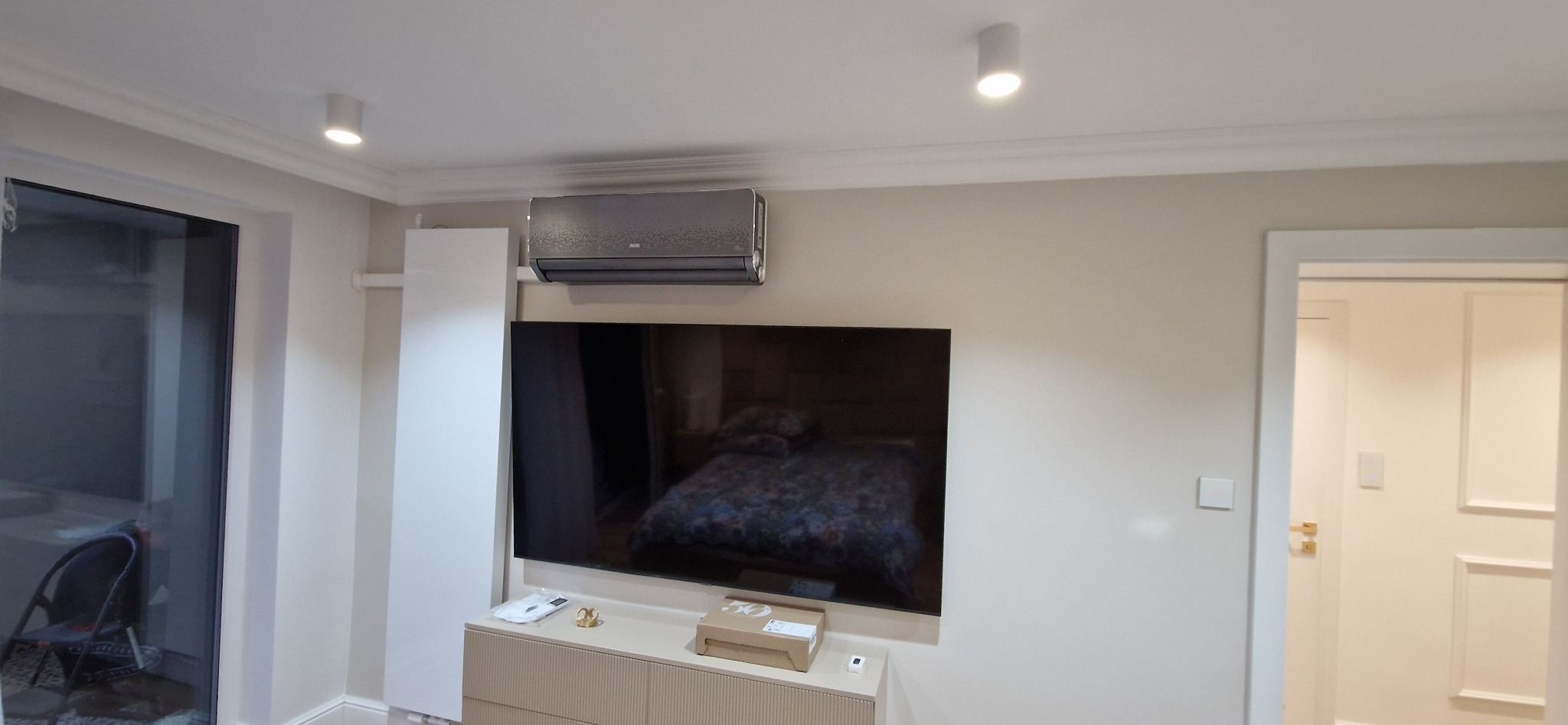 Szary klimatyzator AUX zamontowany nad telewizorem w jasnym salonie z dekoracyjnymi listwami przysufitowymi i punktowym oświetleniem.