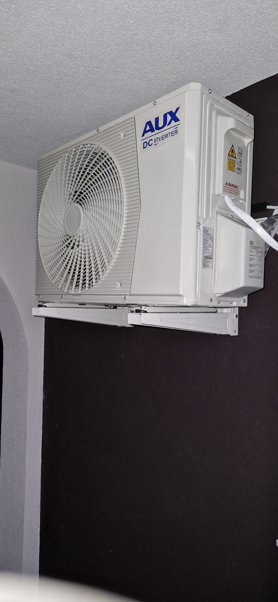 Biała jednostka klimatyzacji AUX DC Inverter zamontowana na ścianie w pomieszczeniu z szarą i brązową ścianą. Widoczne przewody i uchwyty montażowe.