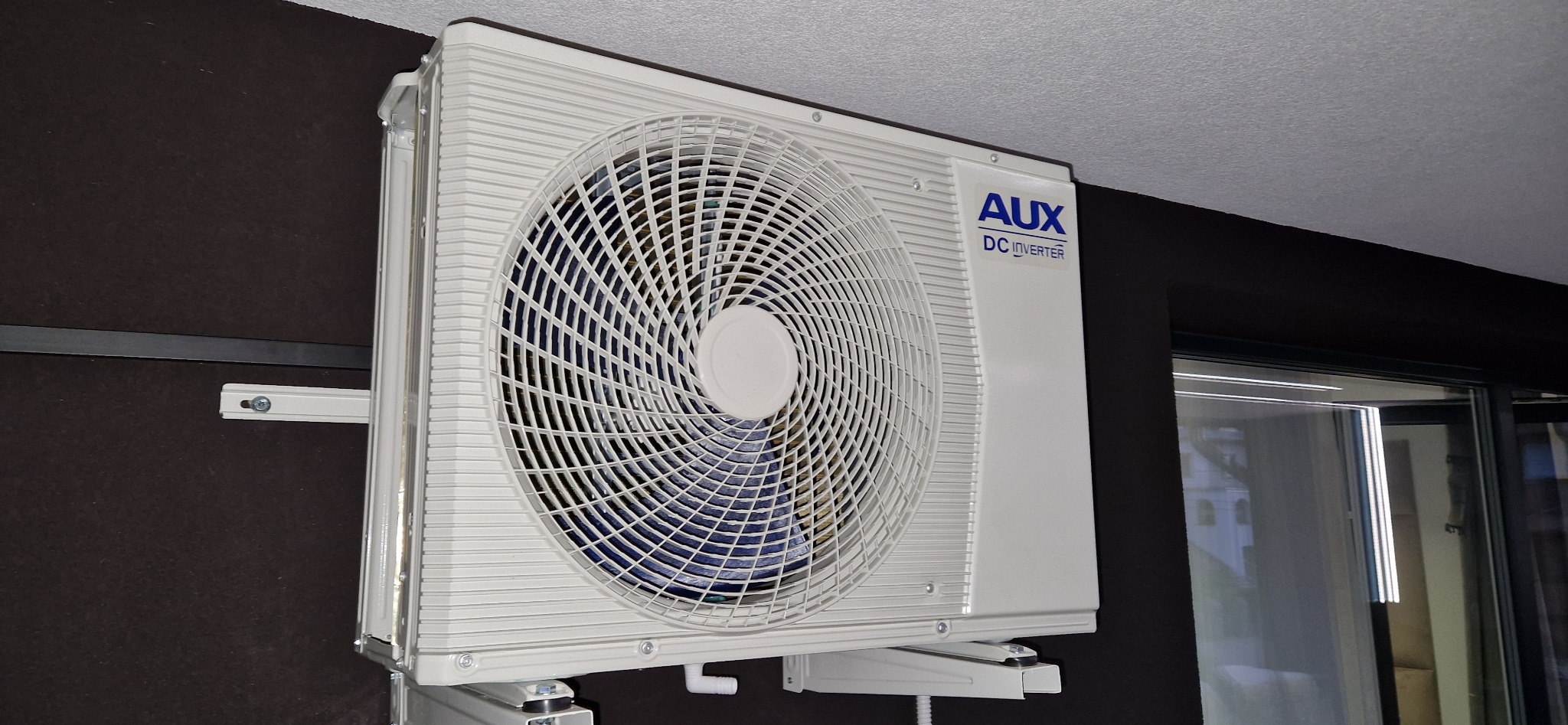 Biała jednostka klimatyzacji AUX DC Inverter zamontowana na brązowej ścianie, widoczna kratka wentylacyjna i fragment okna.