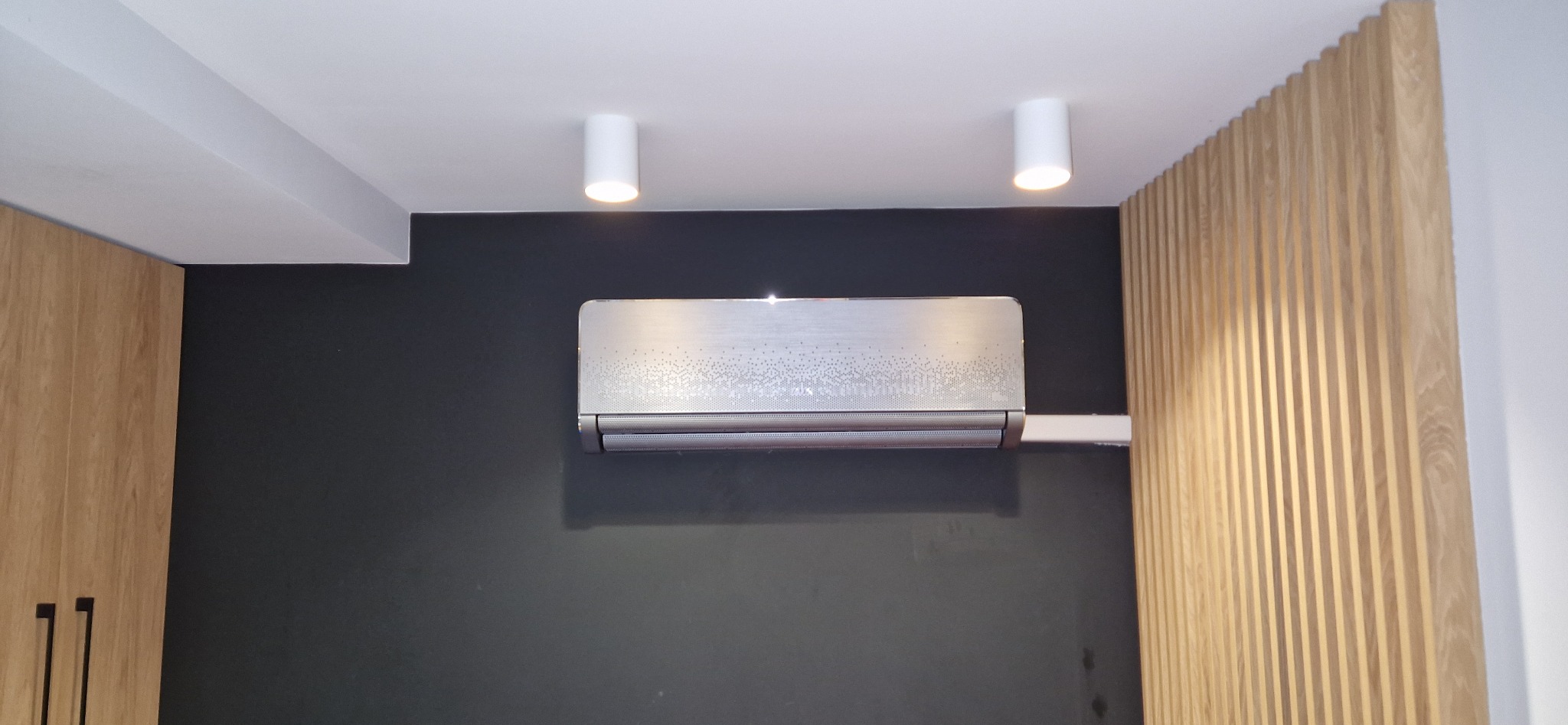 Zamontowany klimatyzator w nowoczesnym wnętrzu w Zambrowie. Srebrny model na czarnej ścianie, oświetlony punktowymi lampami. Elementy dekoracyjne z drewna.