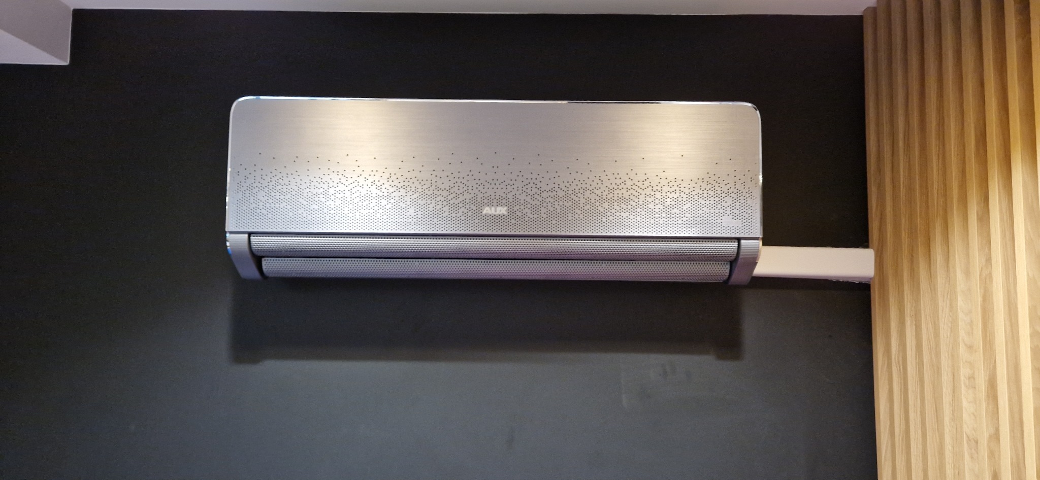 Elegancki, srebrny klimatyzator AUX zamontowany na ciemnej ścianie, z widoczną listwą przypodłogową i drewnianą boazerią po prawej stronie.