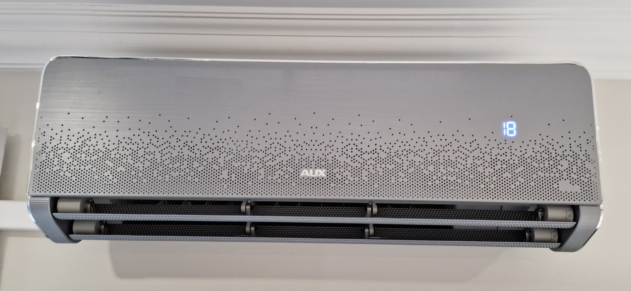 Nowoczesny klimatyzator AUX w odcieniach szarości, z perforowanym panelem i wyświetlaczem temperatury 18, zamontowany na ścianie. Widoczne detale wykończenia i logo.