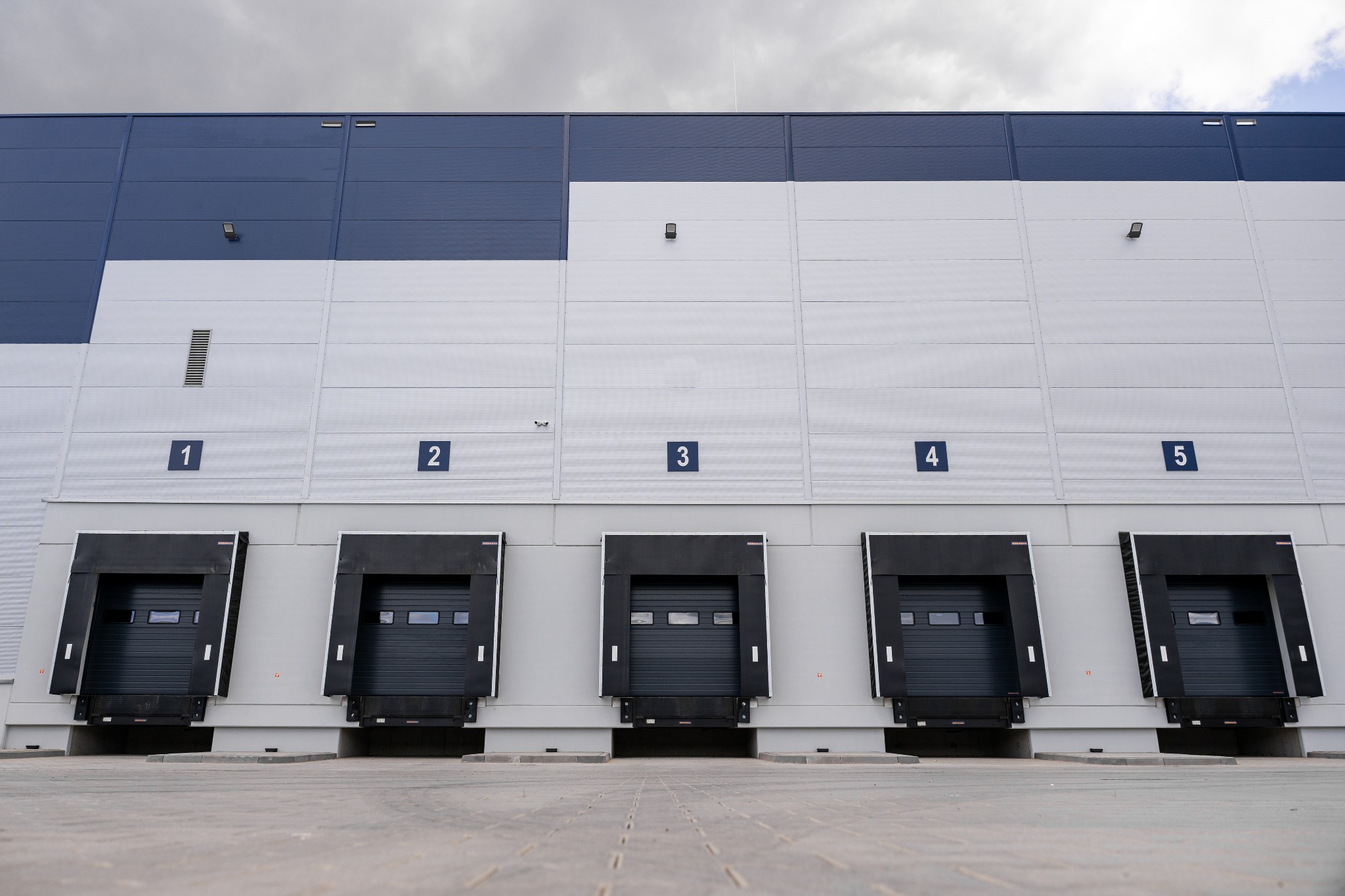 Zewnętrzny widok magazynu z pięcioma rampami załadunkowymi z numeracją od 1 do 5, zamkniętymi czarnymi bramami segmentowymi i szaro-białą elewacją pod pochmurnym niebem.