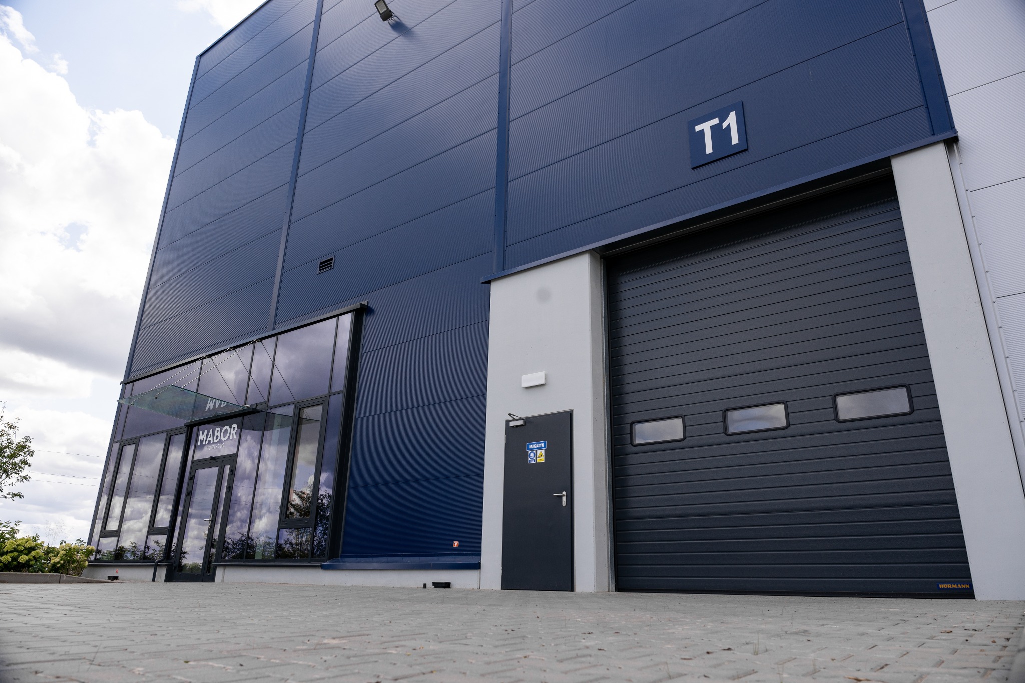 Niebieski budynek magazynowy z szarą bramą segmentową, oznaczony numerem T1, z biurem firmy Mabor i szarym chodnikiem z kostki brukowej.