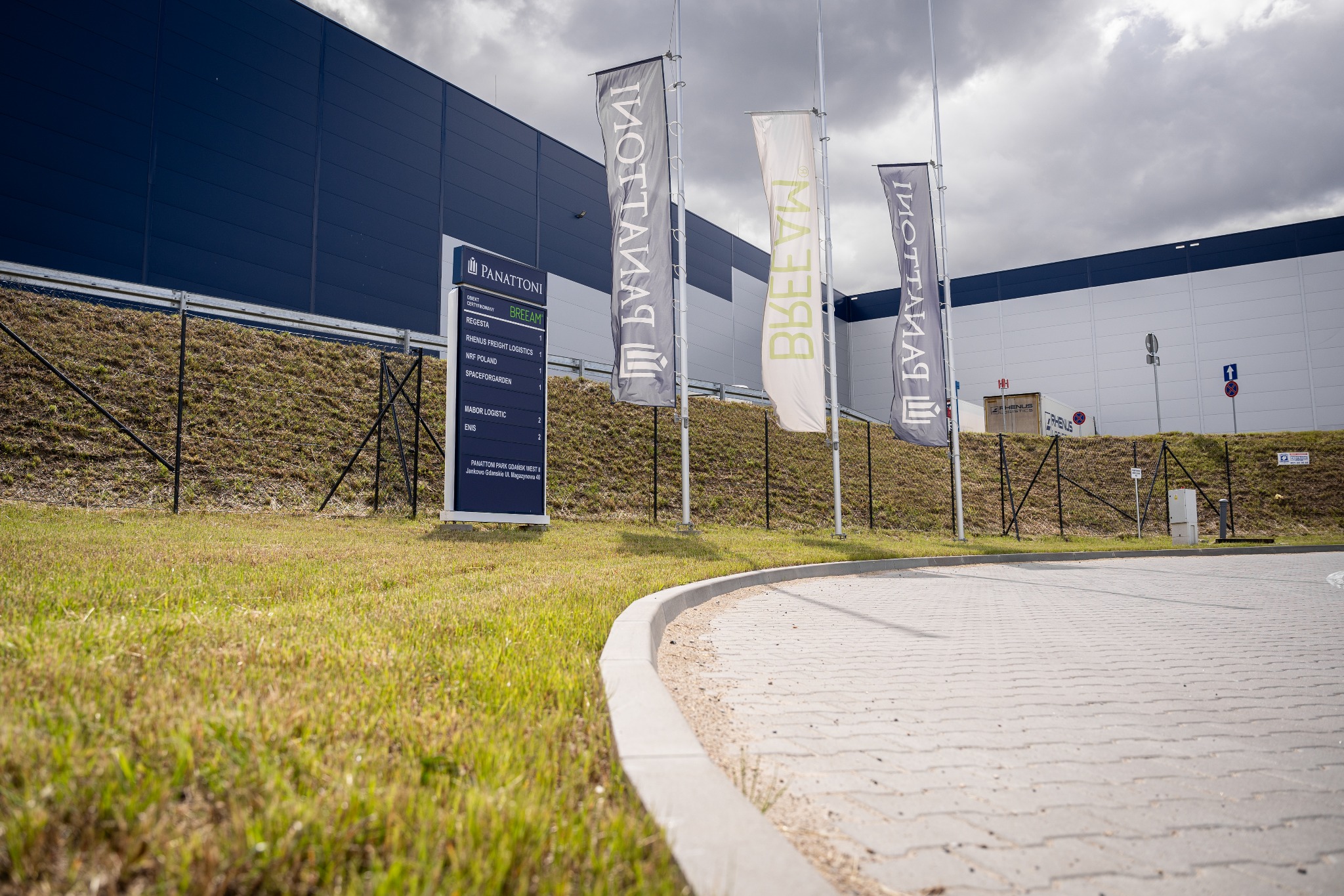 Zewnętrzny widok na nowoczesny magazyn Panattoni Park Gdańsk West z trzema flagami i tablicą informacyjną na tle pochmurnego nieba.