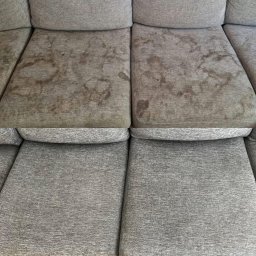MebloPranie LUBIN Pranie tapicerki meblowej , wykładzin , dywanów - Szara sofa z widocznymi plamami na dwóch poduszkach siedziska, kontrastująca z czystymi, dolnymi poduszkami.