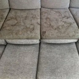 Szara sofa z widocznymi plamami na dwóch poduszkach siedziska, kontrastująca z czystymi, dolnymi poduszkami.