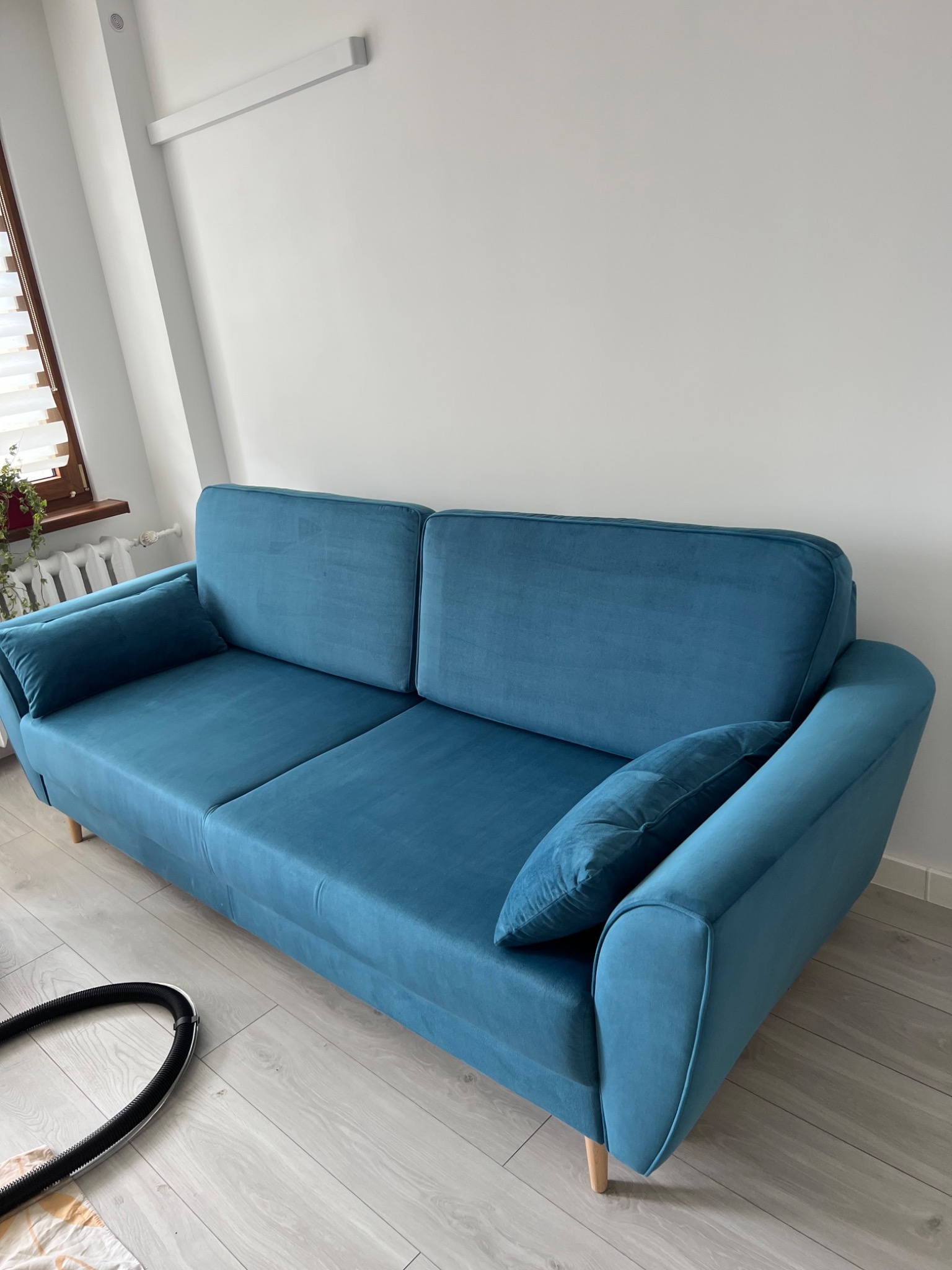 Turkusowa sofa dwuosobowa z poduszką, ustawiona w jasnym wnętrzu z drewnianą podłogą, obok rury od odkurzacza.