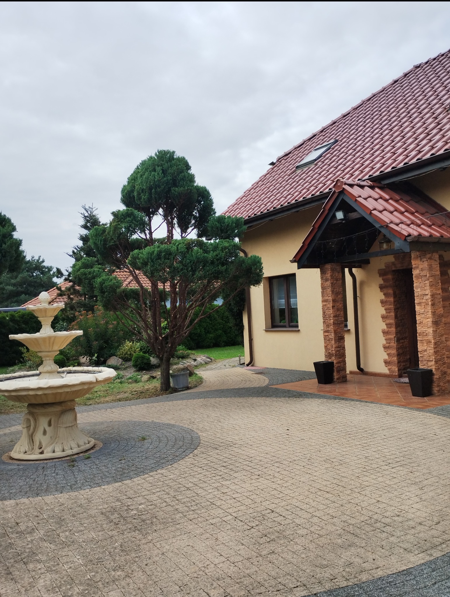 Ogród z fontanną i ozdobnie przyciętym drzewem na tle domu z czerwonym dachem i kolumnami z cegły.