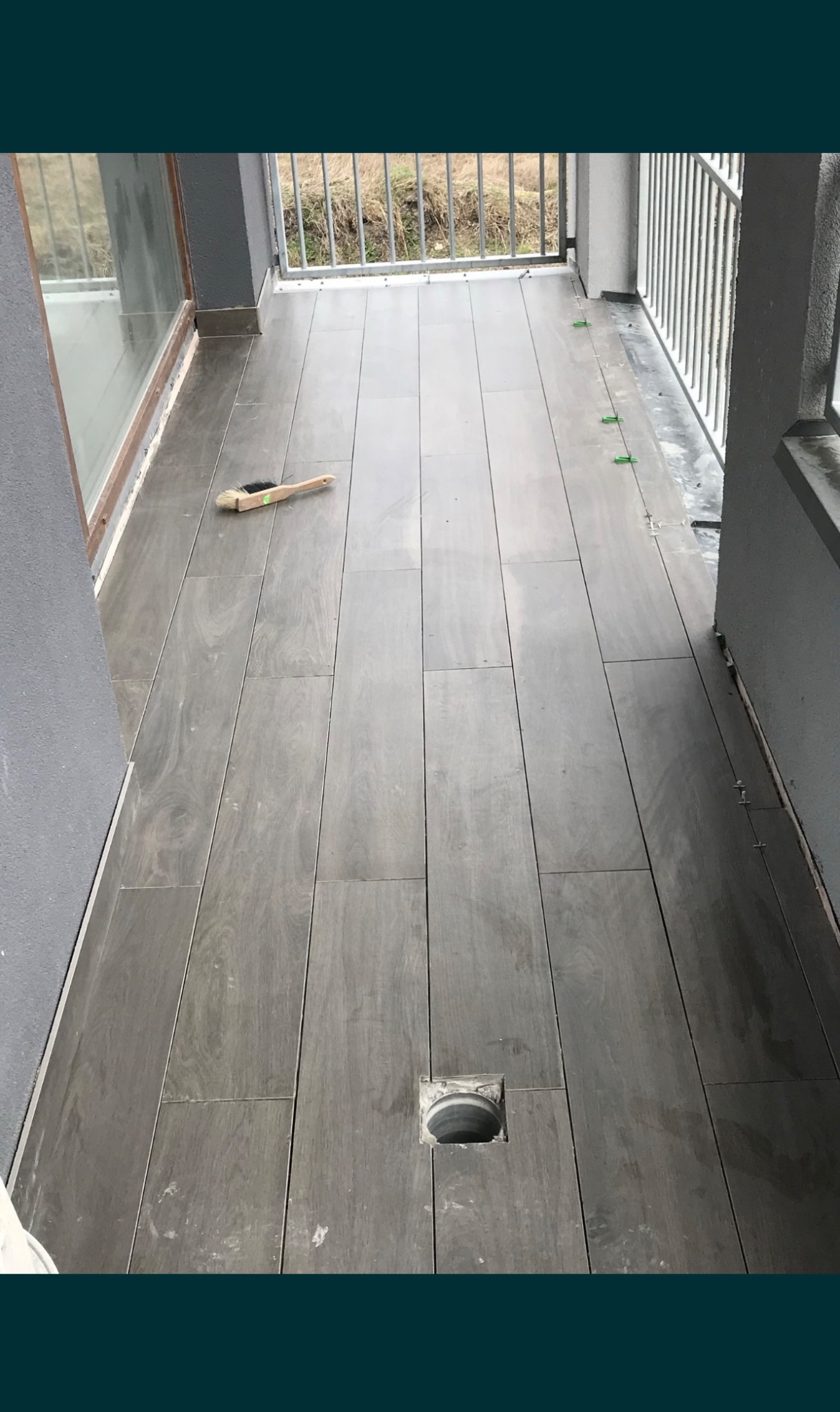 Ułożone szare płytki imitujące drewno na balkonie, widoczny odpływ liniowy, szczotka do zamiatania i plastikowe krzyżyki dystansowe.