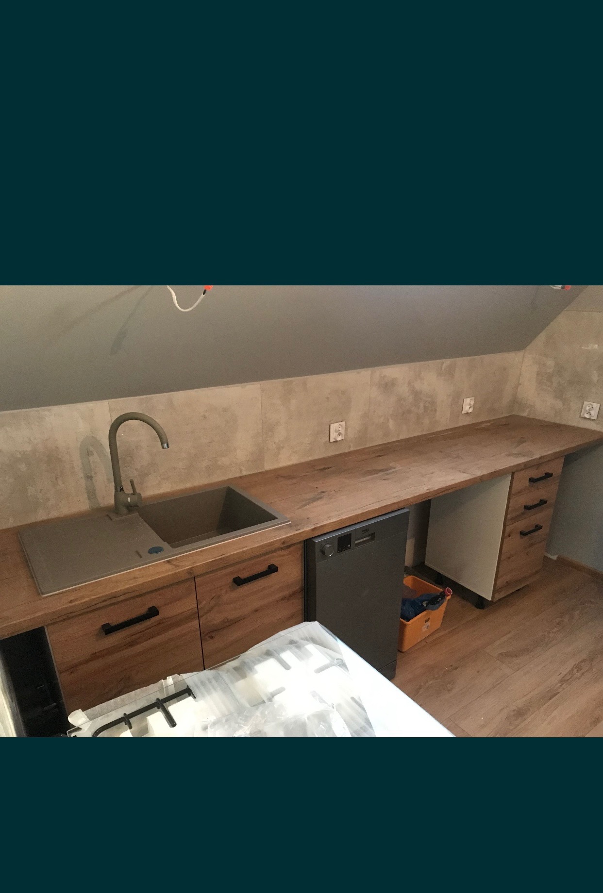 Ukończona zabudowa kuchenna na poddaszu ze zlewozmywakiem, armaturą, blatem z drewnopodobnego laminatu, szarą zmywarką i fragmentem nowej płyty gazowej w folii ochronnej.