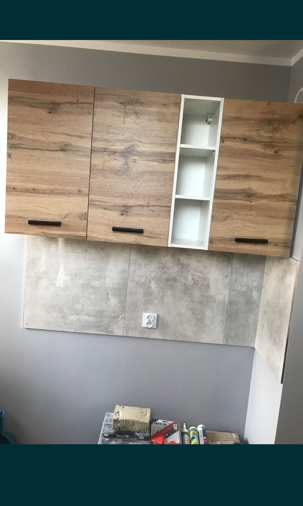 Zamontowane szafki kuchenne z drewnopodobnymi frontami i białym regałem, szare płytki na ścianie, widoczne narzędzia i materiały budowlane na dole zdjęcia.