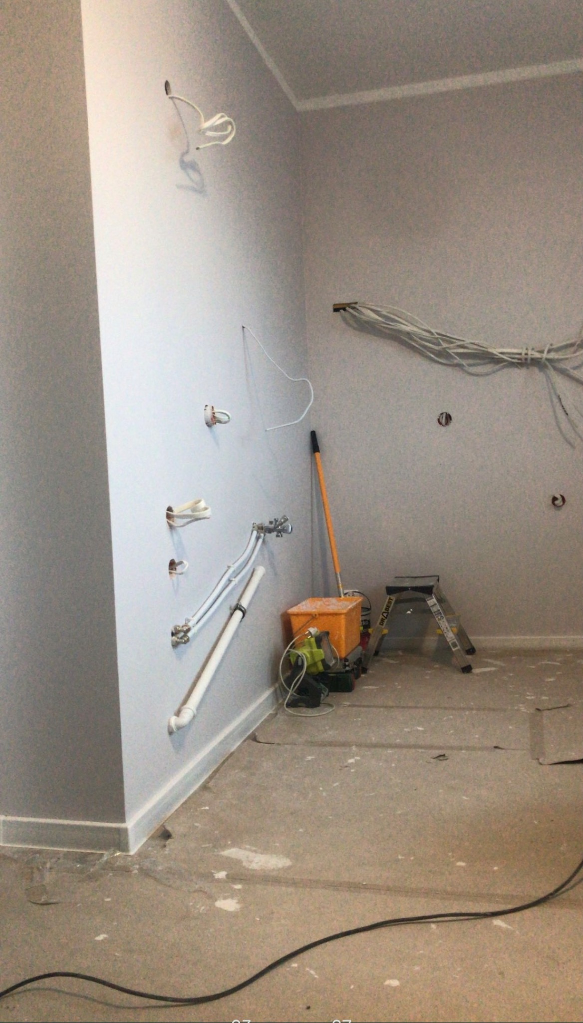 Ściana w szarym pomieszczeniu z wystającymi kablami elektrycznymi i rurami wodnymi, obok stoi drabina i wiadro z narzędziami.