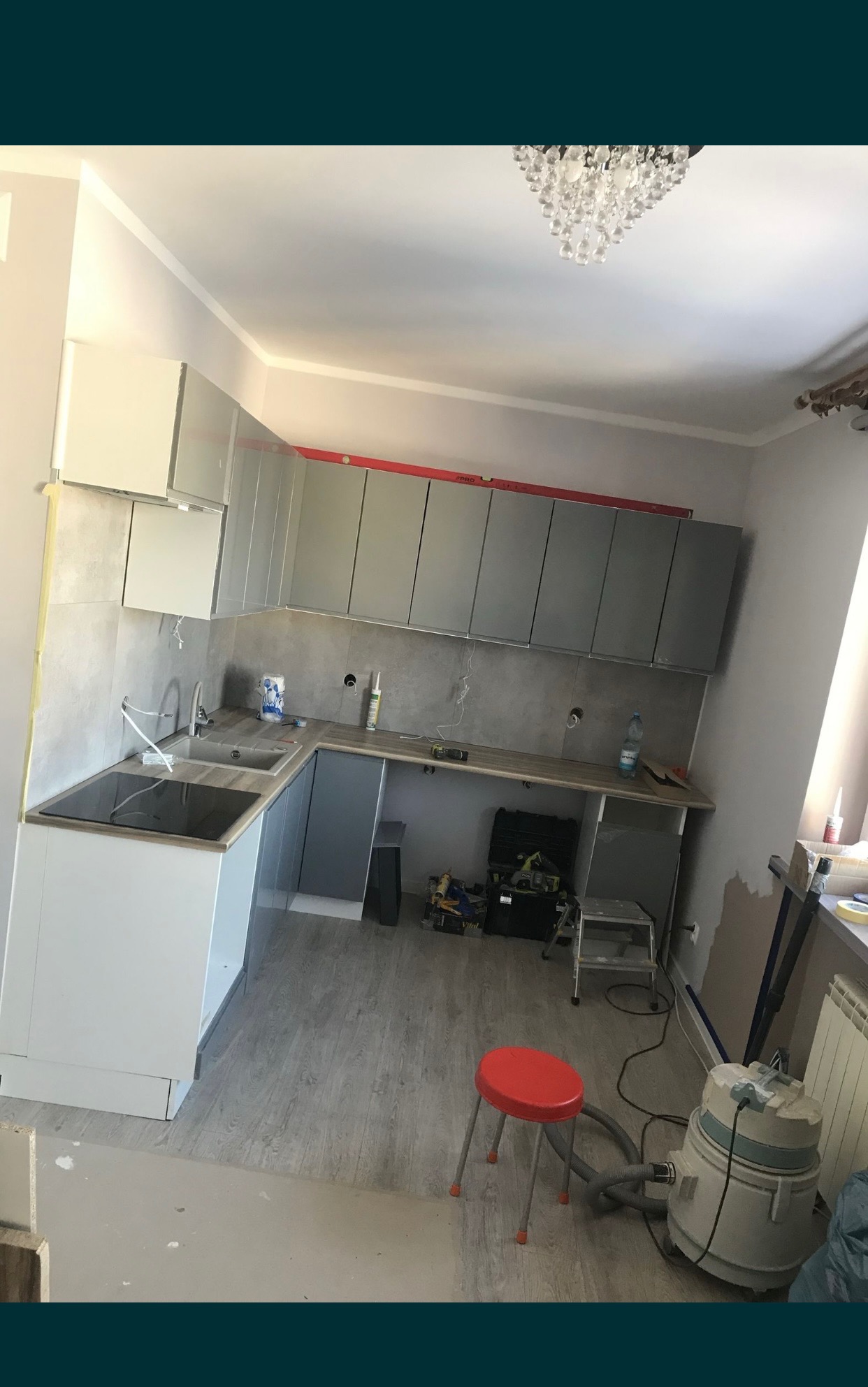Wnętrze kuchni w trakcie montażu: szare szafki wiszące z poziomnicą, białe szafki dolne z czarną płytą indukcyjną, szare płytki na ścianie, widoczne narzędzia, czerwony taboret i odkurzacz...