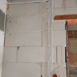 StalLine - Ściana z betonu komórkowego z widocznymi przewodami elektrycznymi prowadzonymi w peszlach, zakończonymi pomarańczowymi puszkami instalacyjnymi, z różowymi oznaczeniami na ścianie.