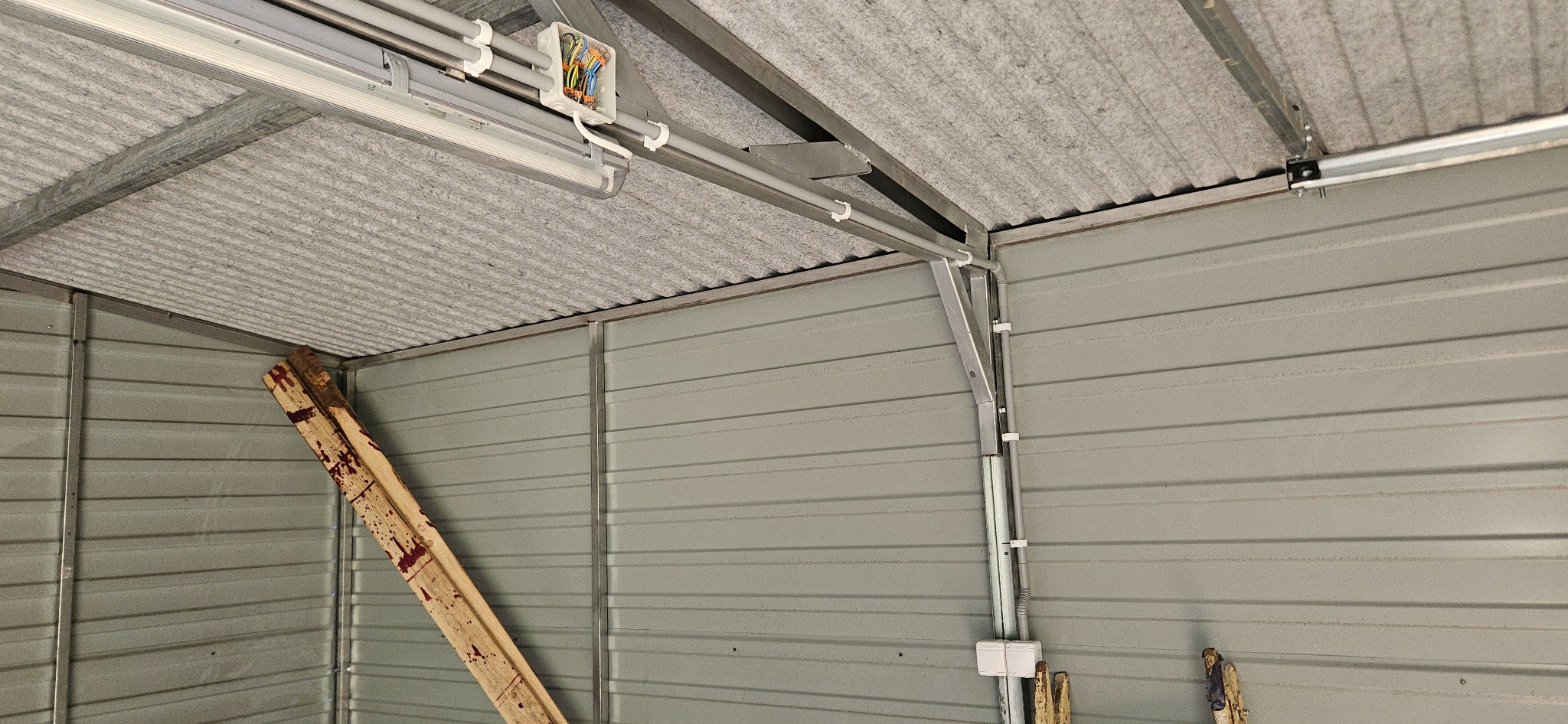 Wnętrze garażu blaszanego w trakcie montażu instalacji elektrycznej: widoczne kable w białych rurkach PCV, puszka elektryczna z kolorowymi przewodami, lampa LED, izolacja dachu i drabina oparta...