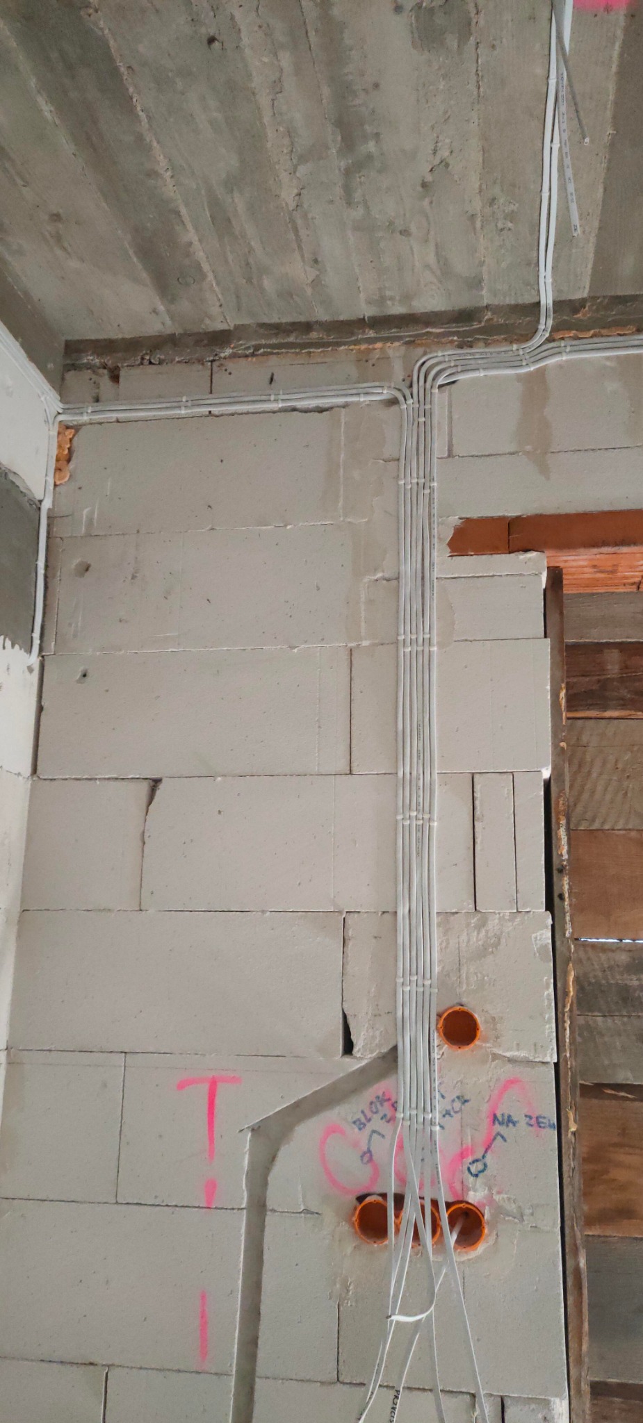 Ściana z betonu komórkowego z widocznymi przewodami elektrycznymi prowadzonymi w peszlach, zakończonymi pomarańczowymi puszkami instalacyjnymi, z różowymi oznaczeniami na ścianie.
