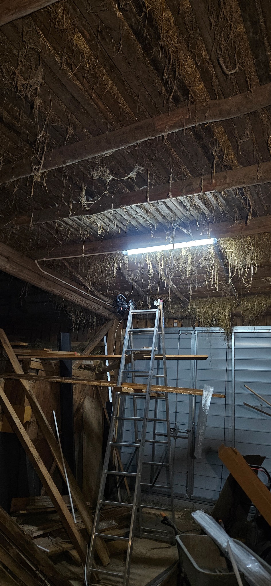 Wnętrze starej hali z drewnianą konstrukcją dachu, gdzie wisi lampa jarzeniowa i stoi aluminiowa drabina, a na niej zamontowane narzędzia.