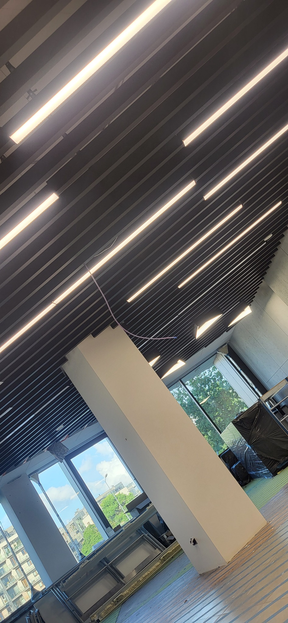 Widok z dołu na sufit z zamontowanym oświetleniem liniowym LED w budynku o nowoczesnej architekturze, widoczny słup konstrukcyjny i okna z widokiem na zieleń miejską.