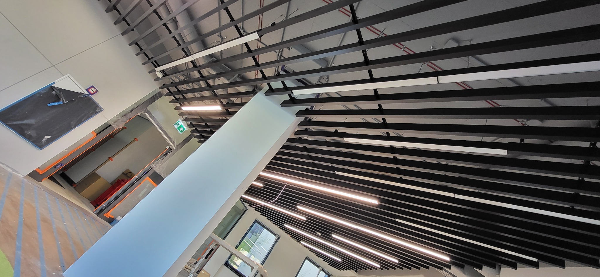 Sufit z czarnymi belkami i zamontowanym liniowym oświetleniem LED, widok z dołu, w tle fragment pomieszczenia w trakcie wykańczania.