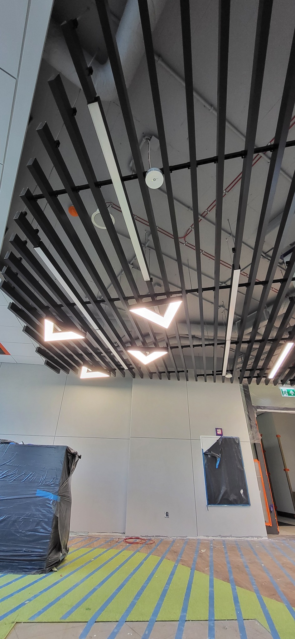 Sufit z czarnymi listwami i nowoczesnym oświetleniem w kształcie litery V, widoczne przewody i instalacje, podłoga zabezpieczona folią i taśmą malarską, ściany w trakcie wykończenia.