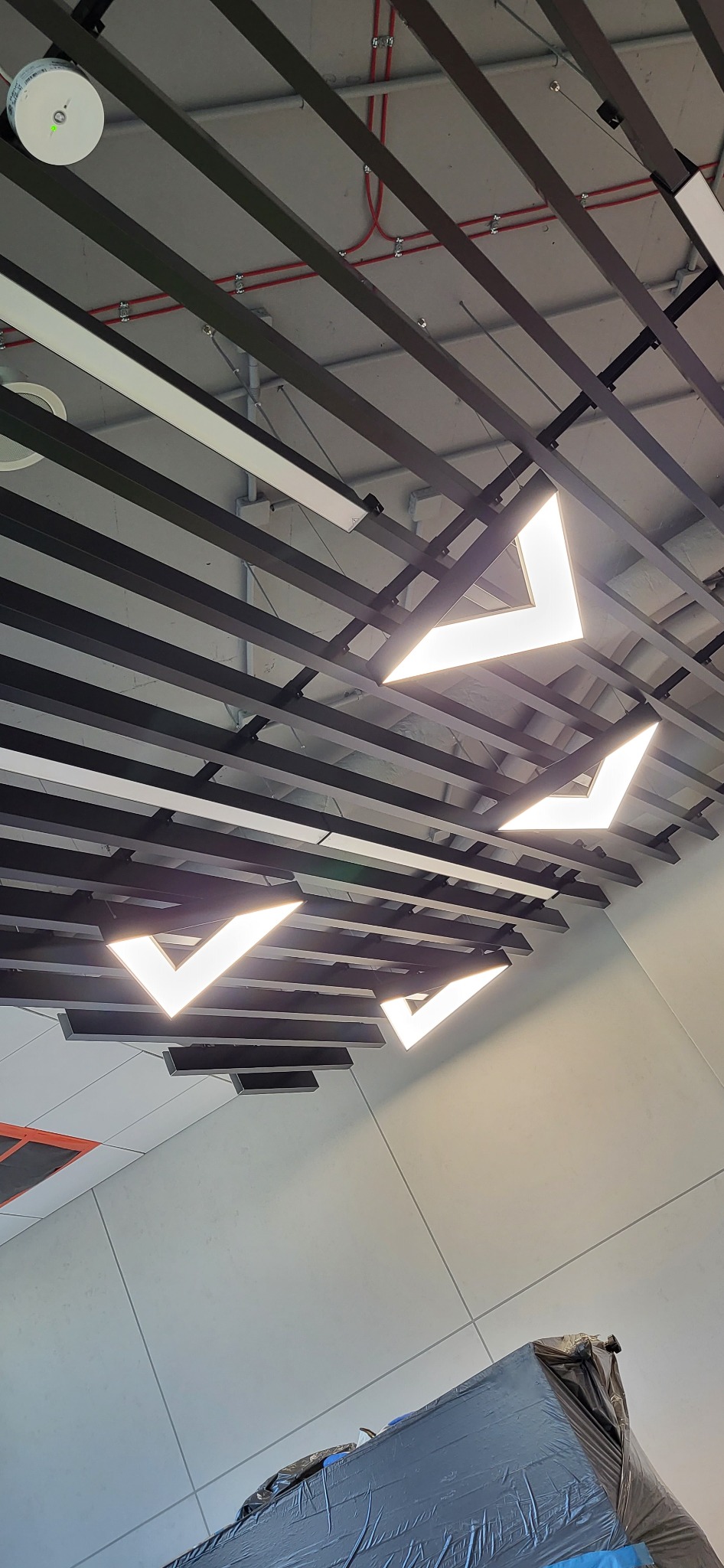 Sufit z nowoczesnym oświetleniem w kształcie litery V, czarne profile konstrukcyjne i widoczne instalacje elektryczne.