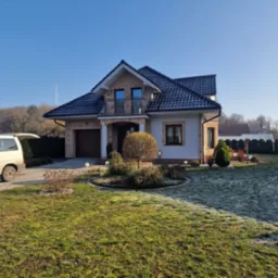 Dwukondygnacyjny dom murowany z ceglaną elewacją, balkonem z kutą balustradą, ciemną dachówką i garażem, otoczony ogrodem z oszronioną trawą w słoneczny poranek.