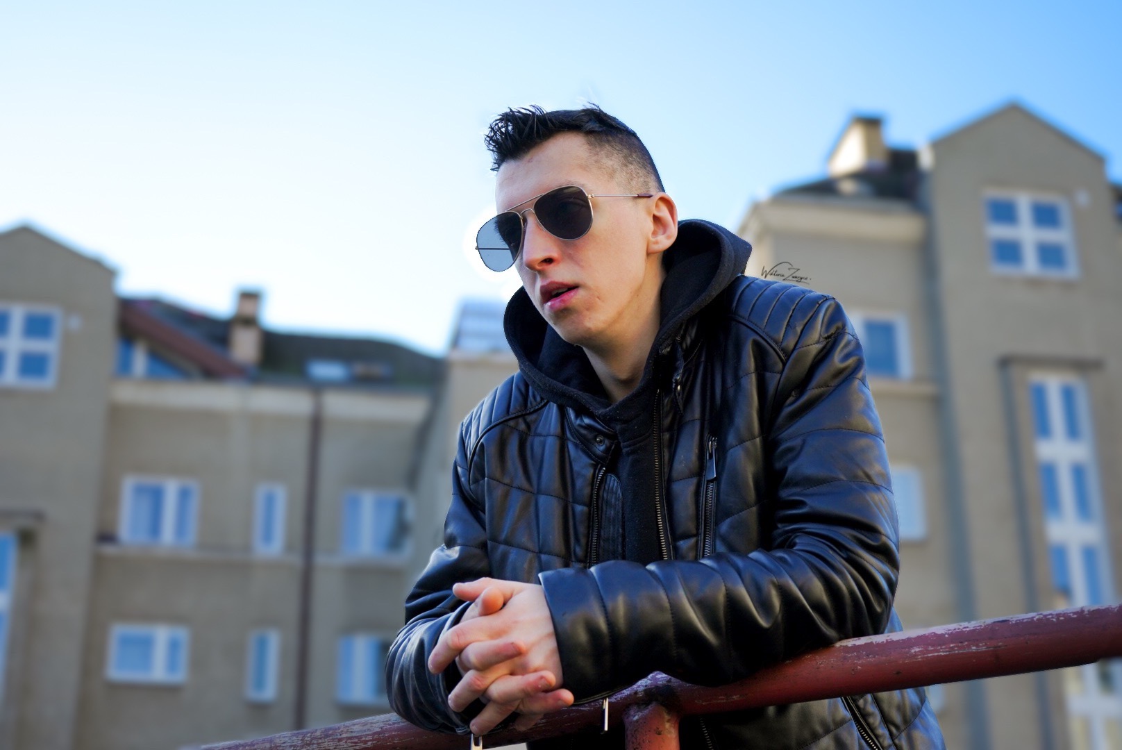 Młody mężczyzna w skórzanej kurtce i okularach przeciwsłonecznych, oparty o czerwoną barierkę na tle jasnego budynku z niebieskimi oknami.