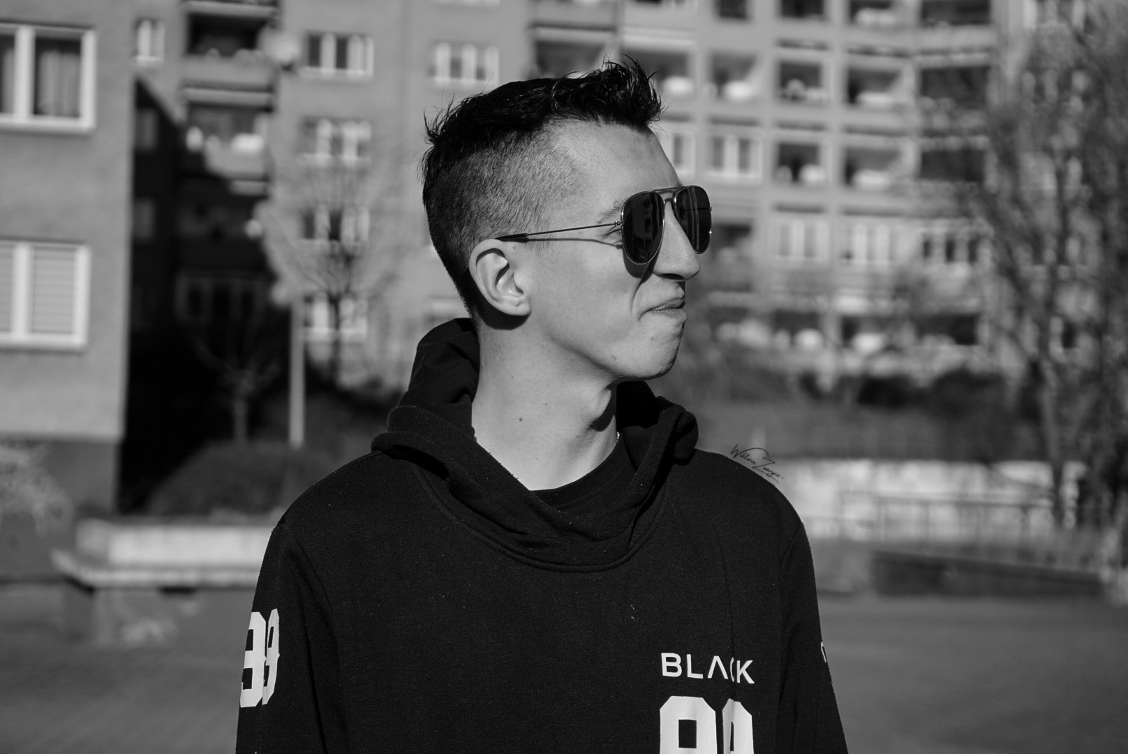 Portret w czerni i bieli: Młody mężczyzna w okularach przeciwsłonecznych i bluzie z kapturem, z budynkiem mieszkalnym w tle.