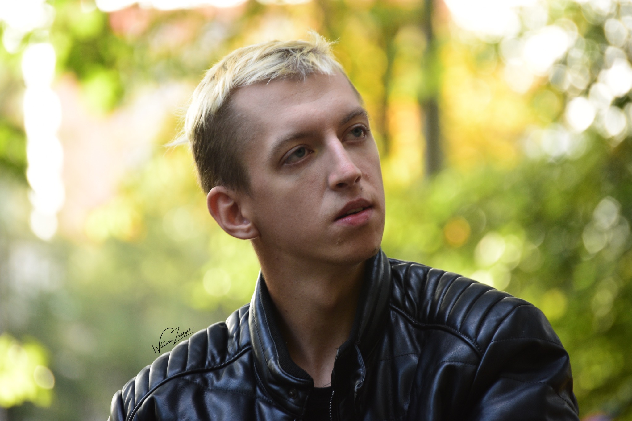 Portret młodego mężczyzny z blond włosami w czarnej skórzanej kurtce, spoglądającego w górę, na tle rozmytych jesiennych liści.