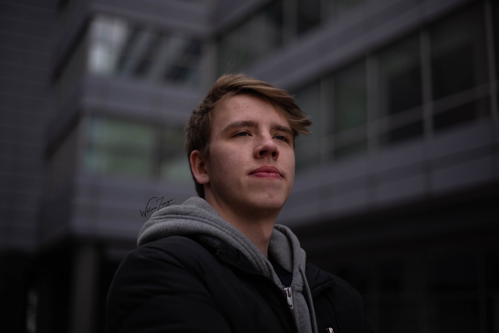 Portret młodego mężczyzny w kurtce i bluzie z kapturem, spoglądającego w górę, na tle rozmytego, nowoczesnego budynku biurowego.