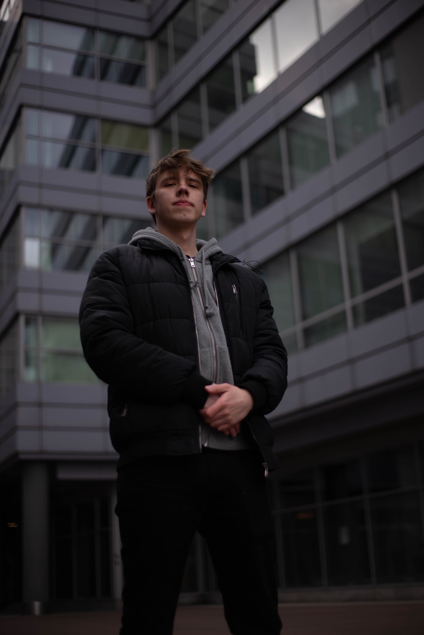 Młody mężczyzna w czarnej kurtce i szarej bluzie z kapturem pozuje na tle nowoczesnego, szklanego budynku o szarych ramach okiennych.