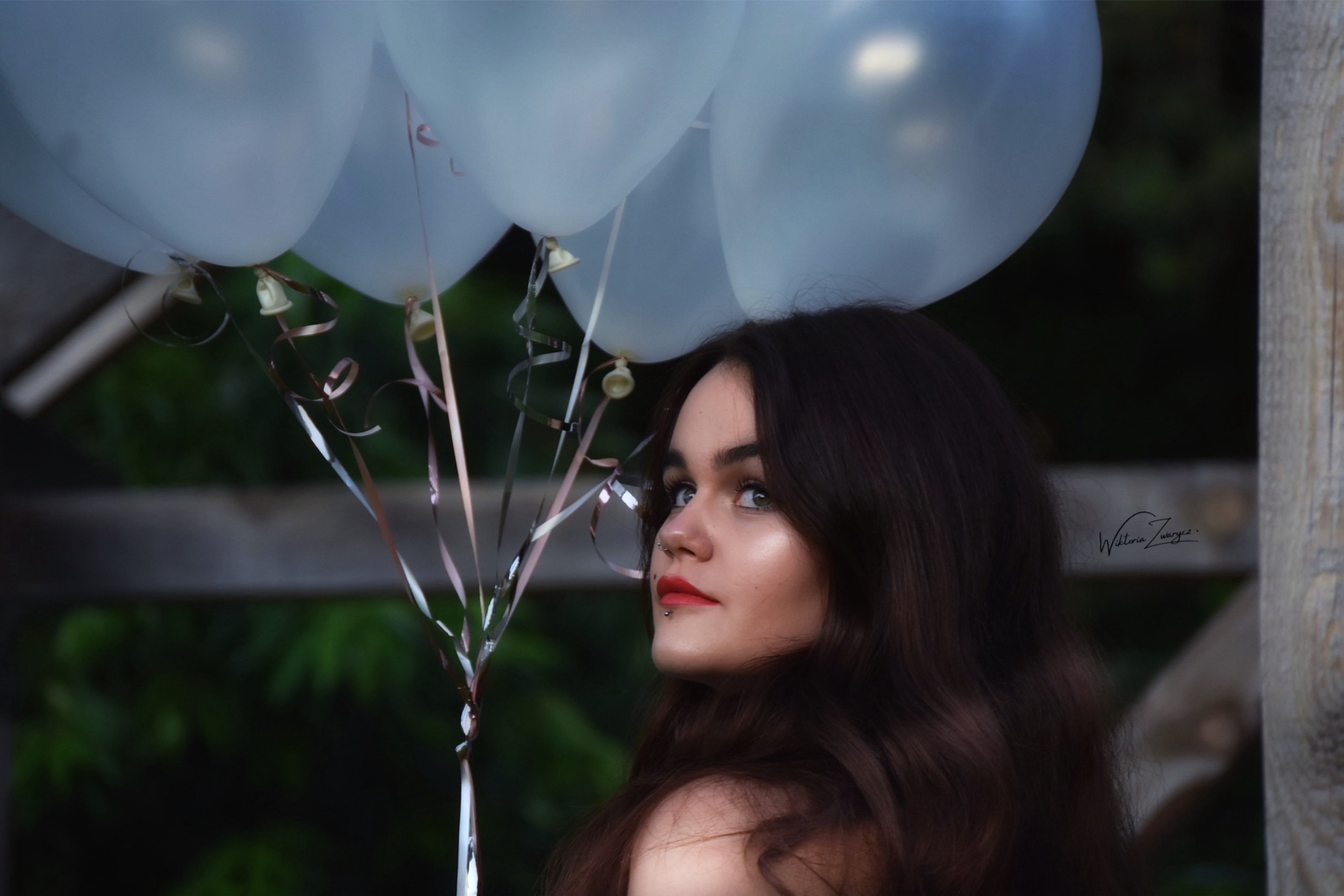 Portret kobiety z balonami w Szczecinie, spoglądającej w bok. Ciemne włosy, czerwone usta, piercing. W tle drewniana konstrukcja i zieleń.