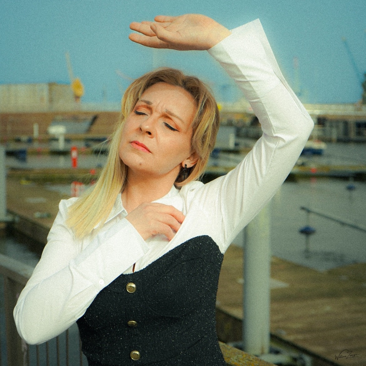 Portret kobiety w białej koszuli i czarnym gorsecie na tle portu, z ręką uniesioną nad głową, chroniącą oczy przed słońcem.