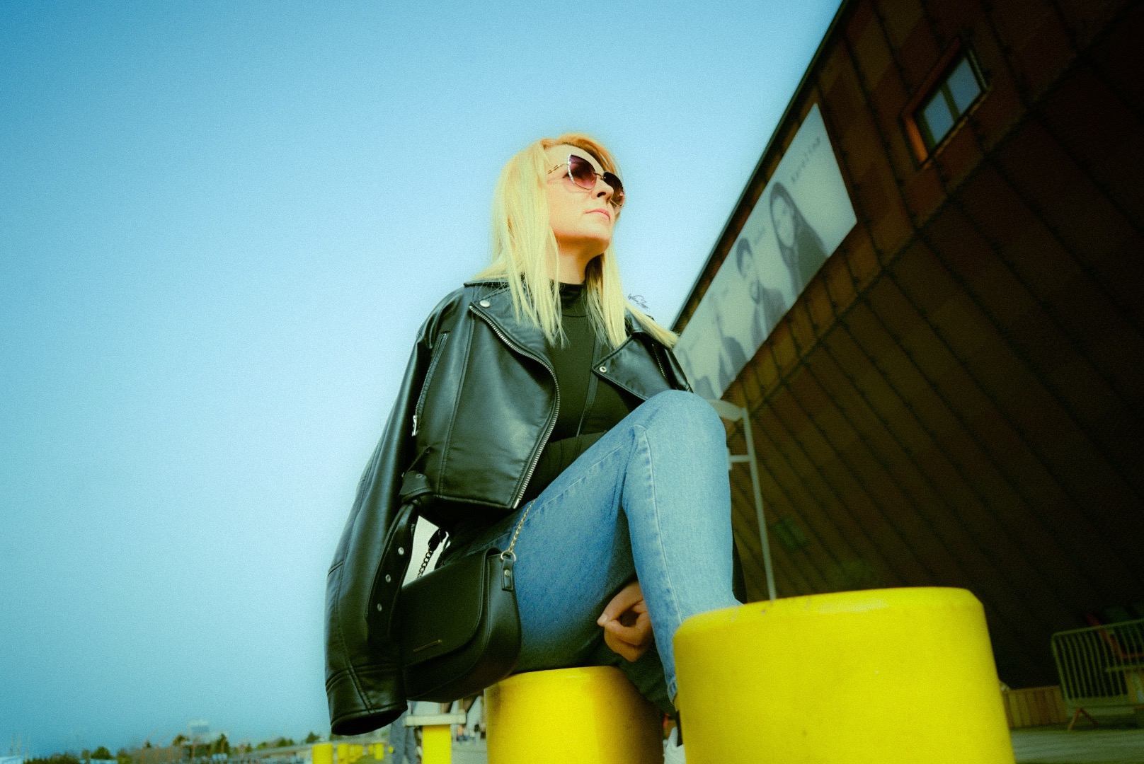 Kobieta z blond włosami w okularach przeciwsłonecznych, czarnej skórzanej kurtce i dżinsach siedzi na żółtym słupku na tle błękitnego nieba i budynku z reklamą.