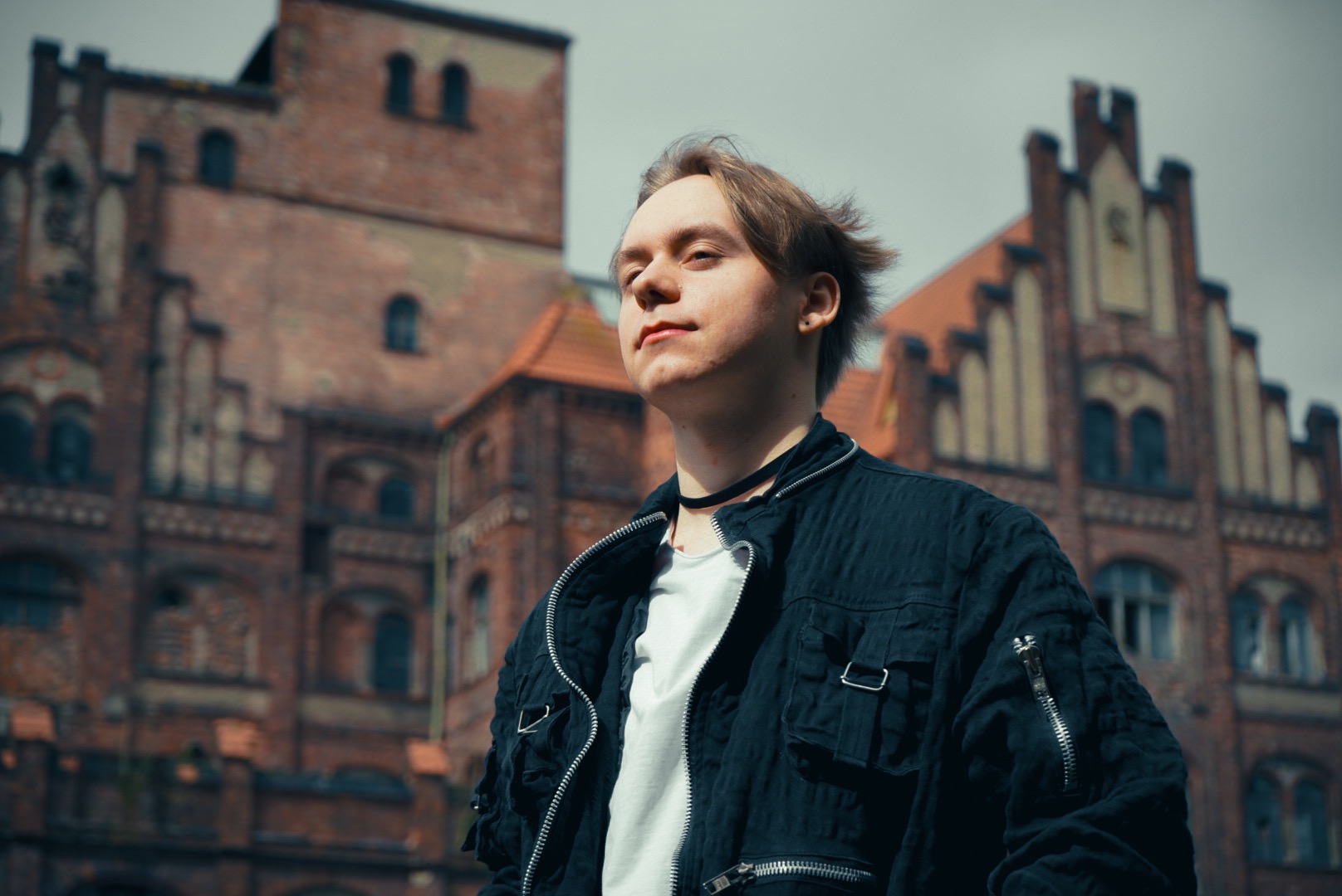 Portret młodego mężczyzny w czarnej kurtce na tle ceglanego budynku z wieżyczkami, z wiatrem we włosach i chokerem na szyi.